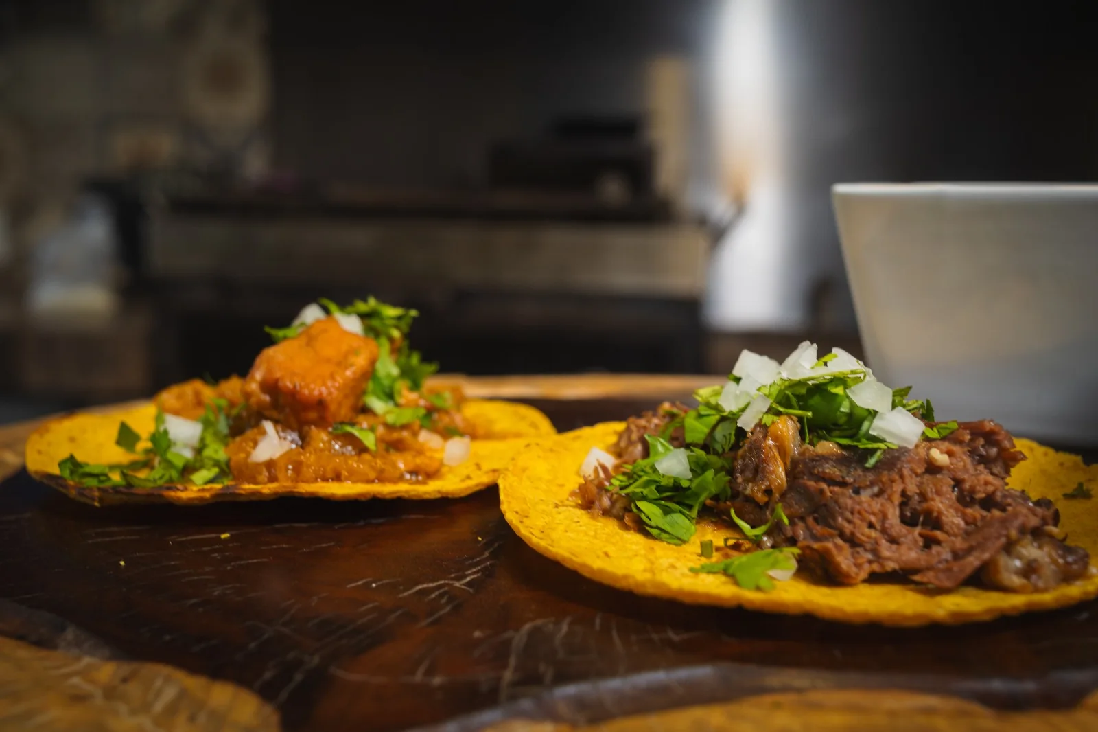 Dos tacos recién servidos sobre una tabla de madera, uno de carne deshebrada y otro de cerdo en salsa roja, con cebolla y cilantro.