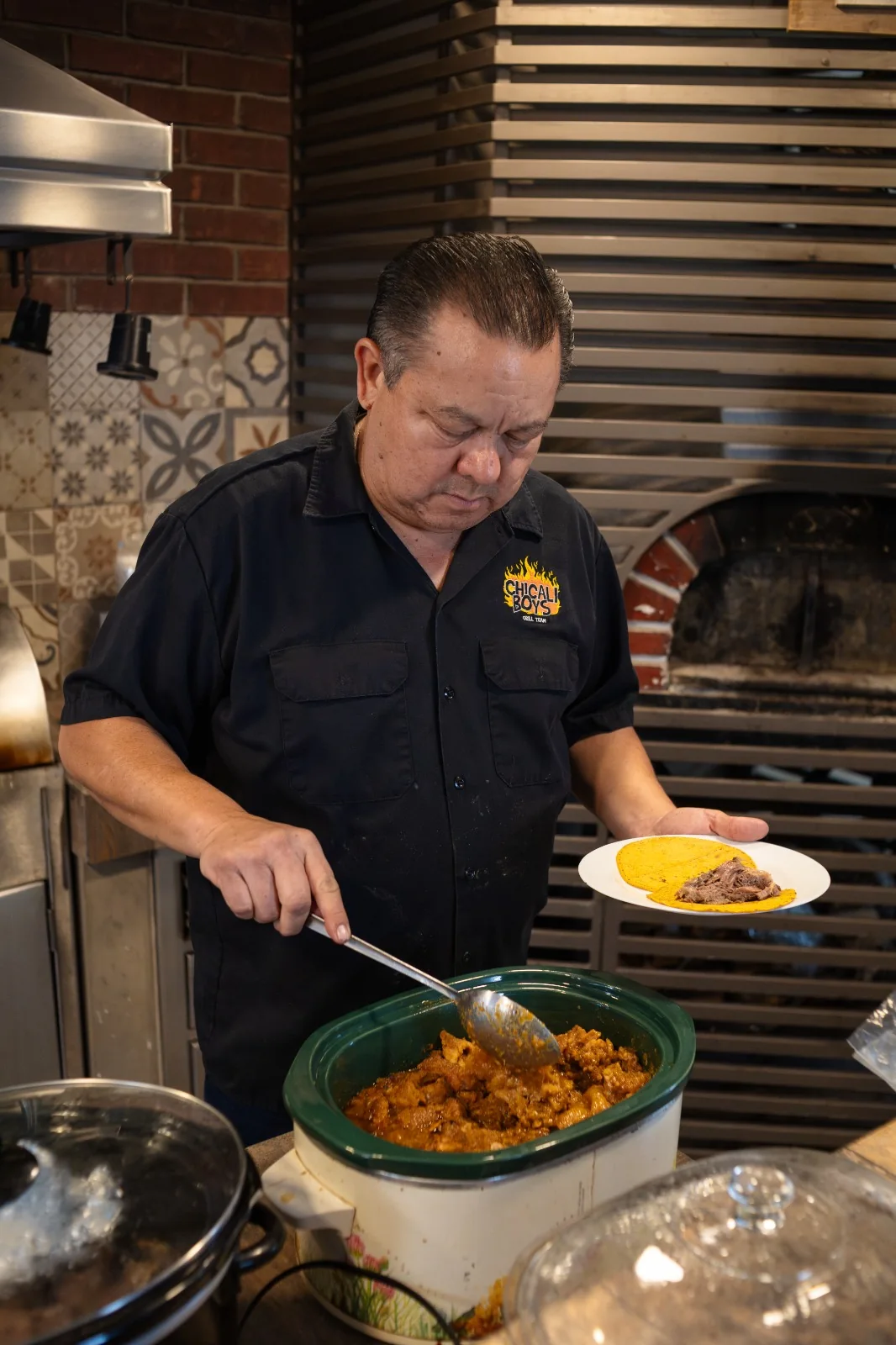 Integrante de Chicali Boys, con camisa del negocio, sirve chicharrón de la olla mientras sostiene un plato con tortillas.