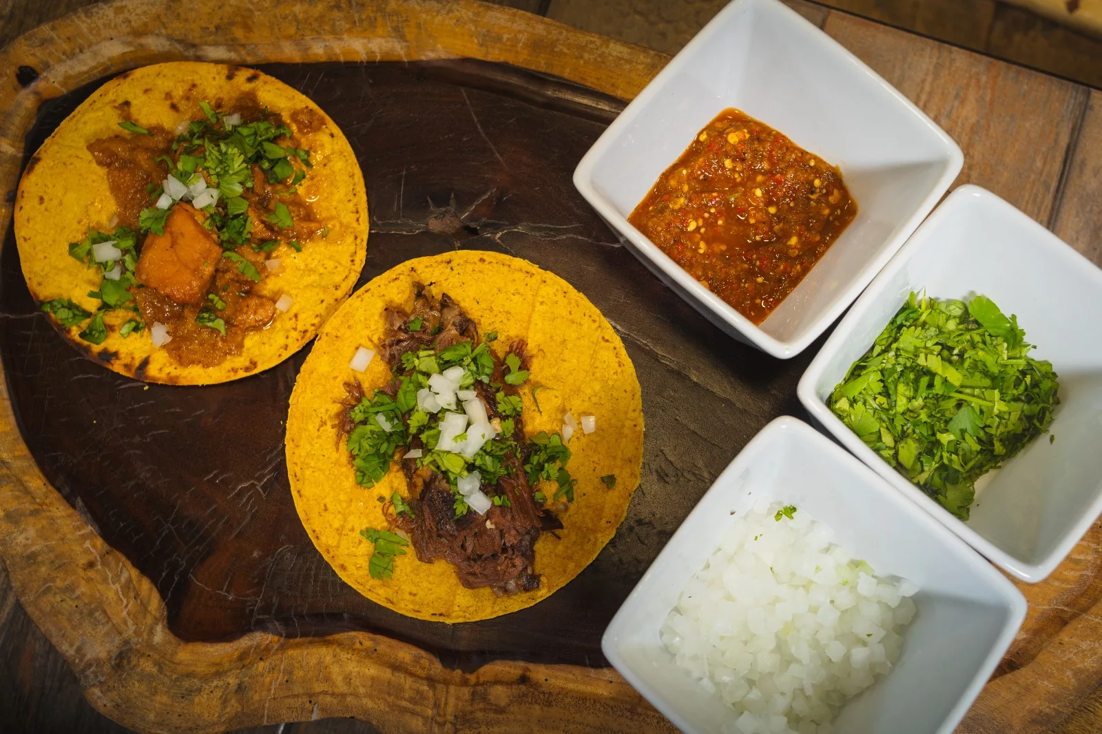 Vista desde arriba de dos tacos sobre una tabla de madera, junto a tazones con salsa roja, cilantro y cebolla picada.
